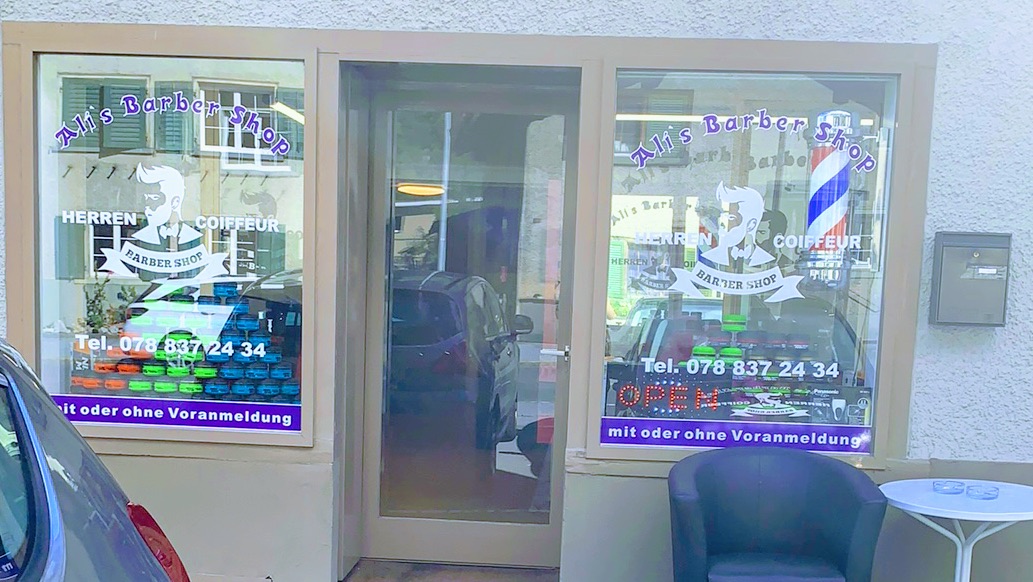 Schaufenster von Ali's Barber Shop mit Logo, Barberstange und Telefonnummer, davor ein Sessel